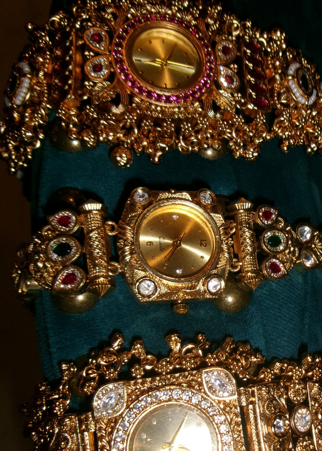 Three ornate gold-tone watch bracelets displayed side by side on a teal velvet surface against a black background. The watches feature intricate filigree bands, crystal embellishments, and colorful gemstone accents in red, green, and white.