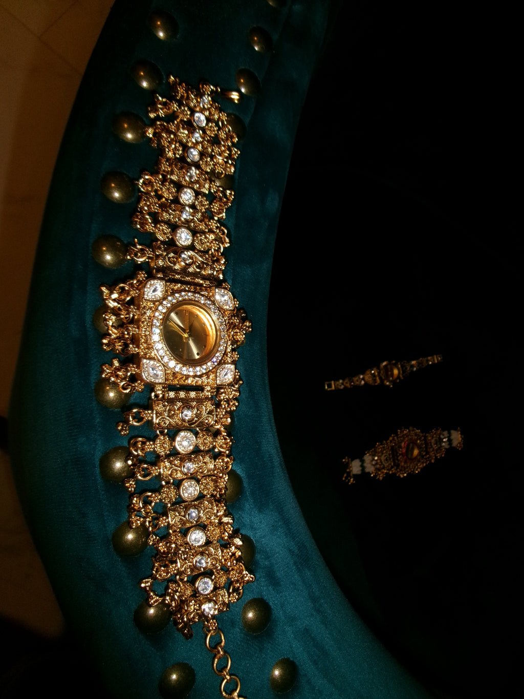 An ornate gold-toned traditional Indian style necklace with intricate jewelry details, clear stones, and gold-toned pearls, displayed on a dark teal velvet surface against a black background.