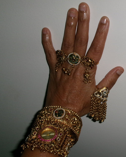 A hand wearing multiple ornate gold-toned rings and a matching wide bracelet, decorated with green, pink, and white stones, against a plain light gray background.