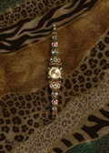 A gold-toned bracelet watch with a round bezel and multiple colorful rhinestone clusters, displayed vertically on a patterned fabric background of leopard print, zebra stripes, and brown tones.