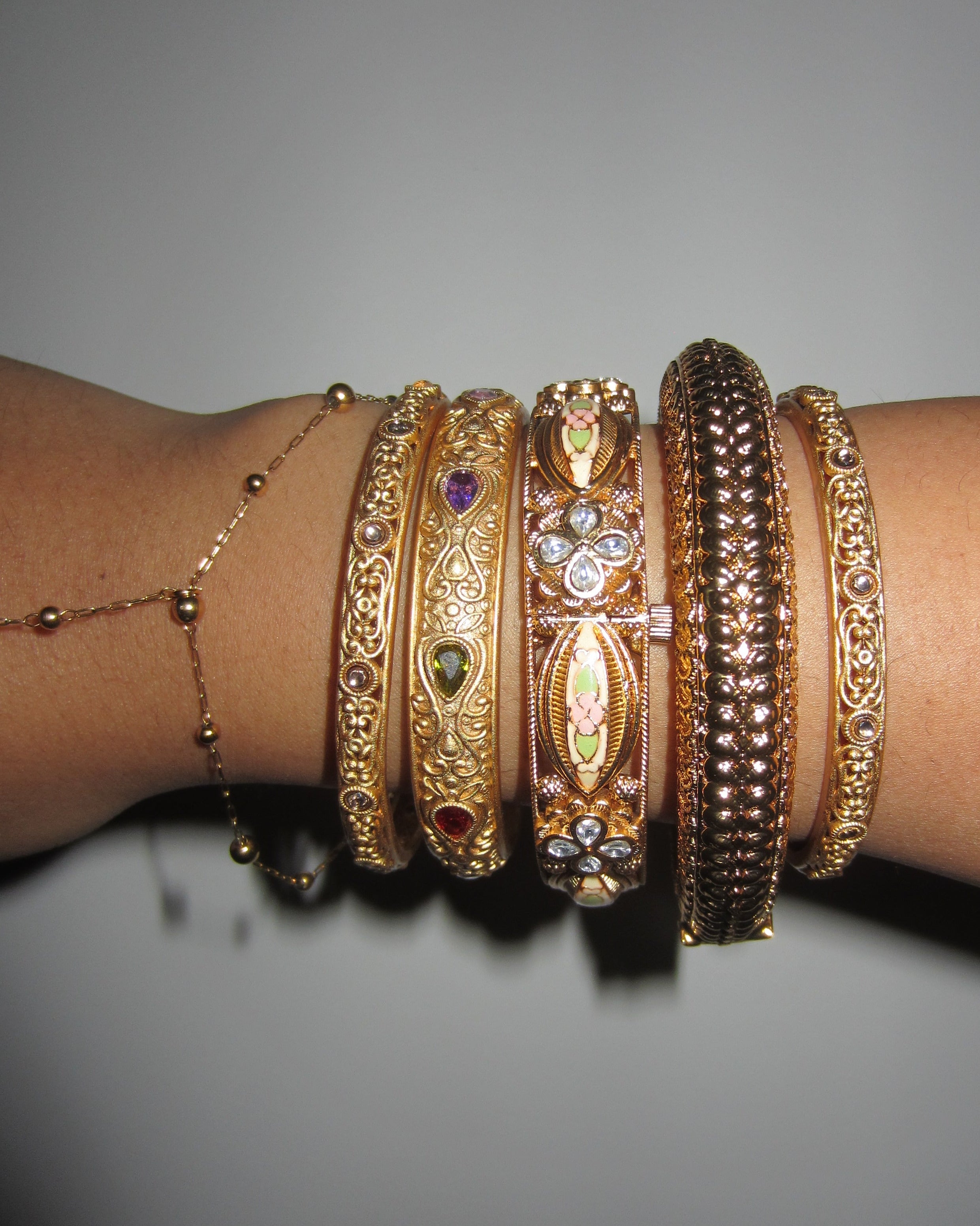 Close-up of a wrist wearing multiple gold bangles with intricate designs on a gray background