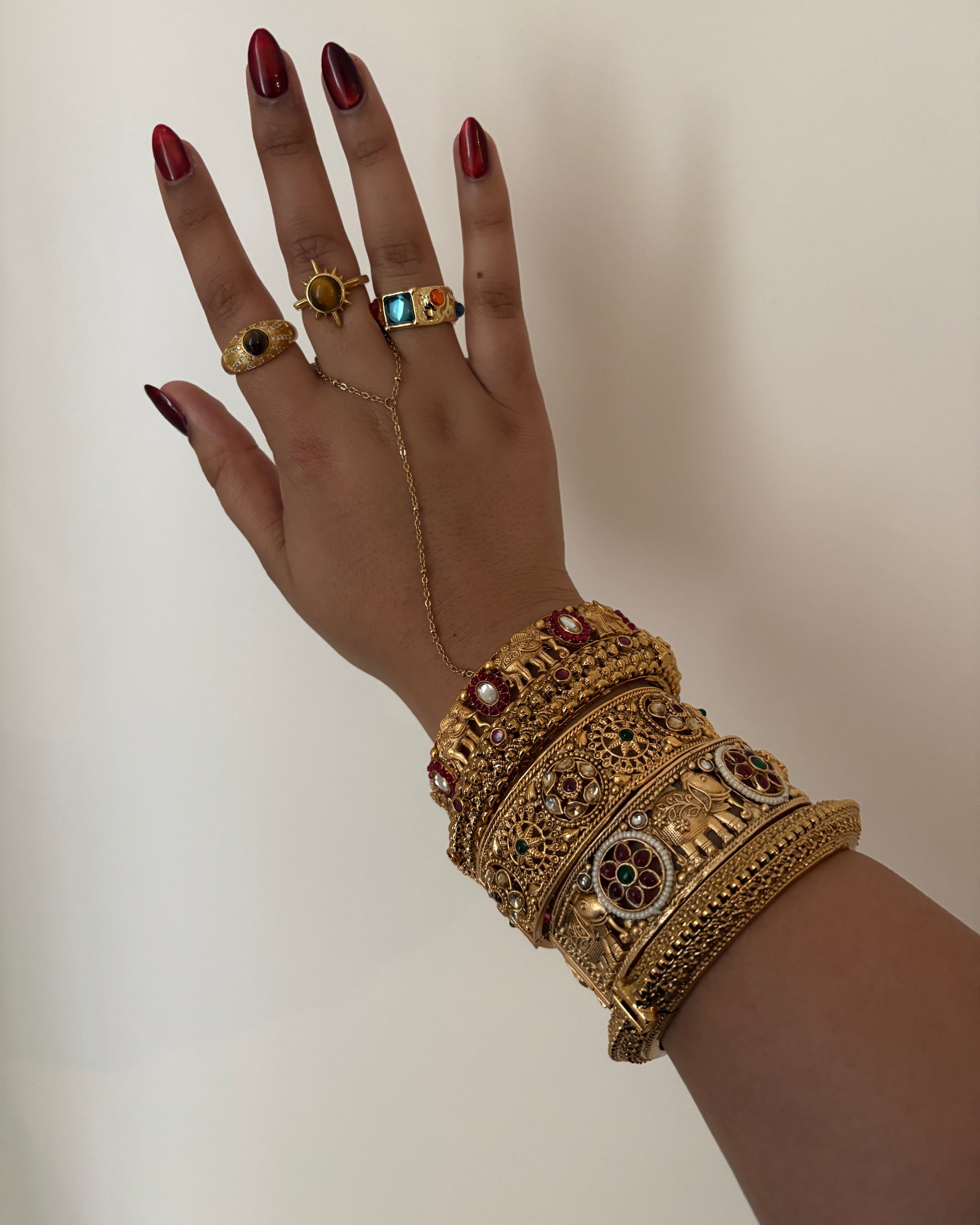 Hand wearing gold jewelry with intricate designs on a neutral background