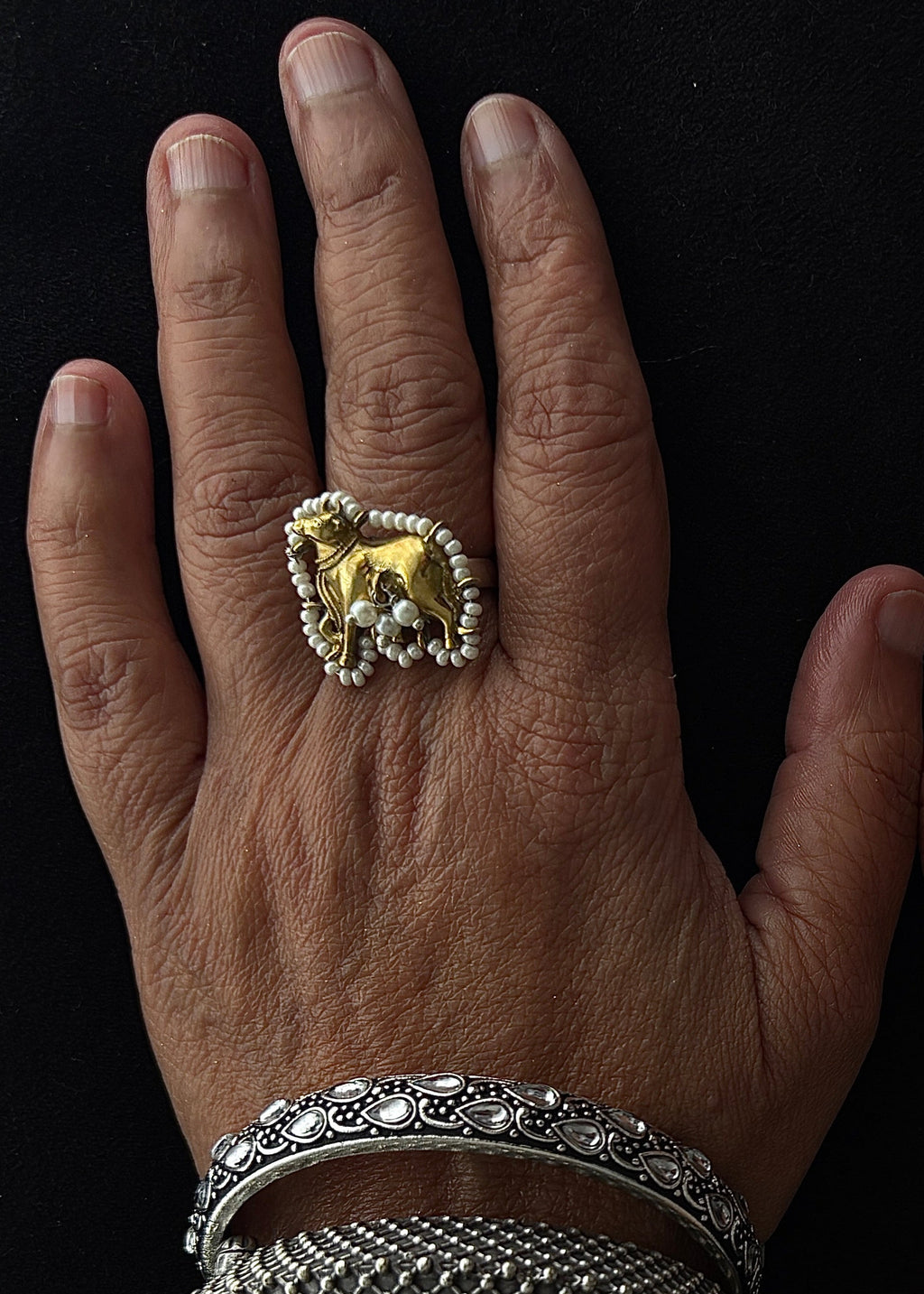 A close-up of a person's hands and wrists adorned with multiple intricate silver-toned traditional bangles and a large gold-toned ring against a black background.