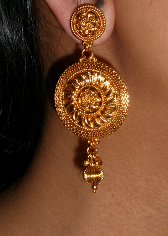 A close-up side profile of a woman wearing a large, ornate gold-toned traditional dangle earring with intricate filigree detailing.