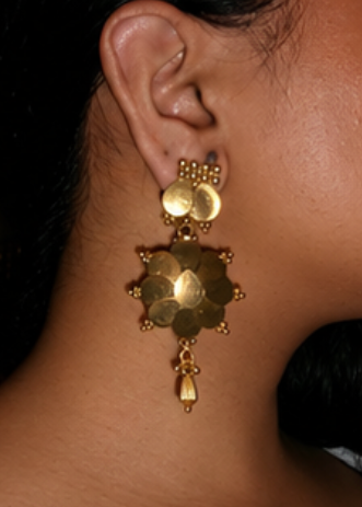 A close-up side profile of a woman wearing a large, ornate gold-toned dangle earring with layered circular and geometric elements.