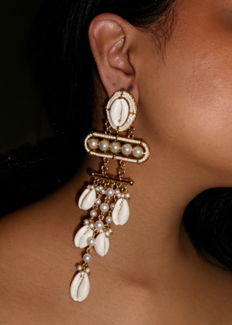 A close-up of a model wearing a large, ornate gold and white dangle earring featuring cowrie shells and pearls.