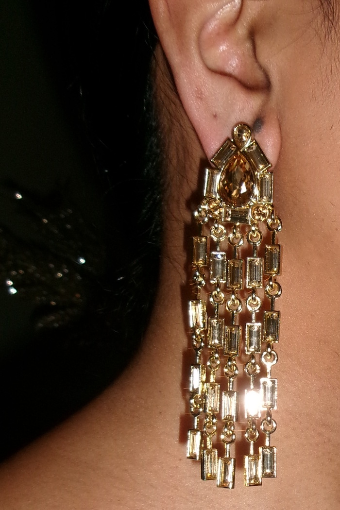 A close-up of a person's ear wearing a long, dangling gold statement earring with multiple cascading rectangular and teardrop-shaped clear stones.